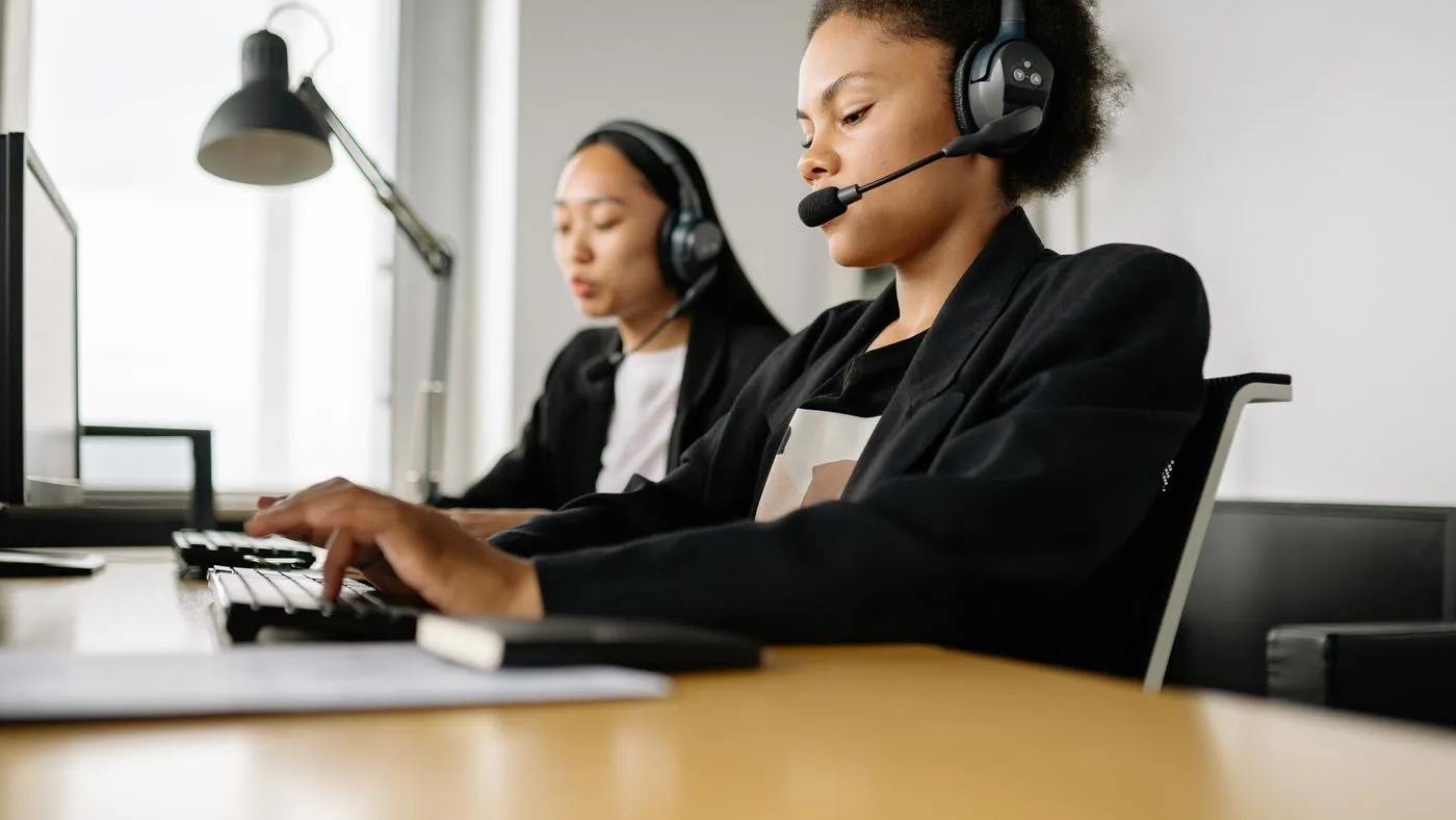 Two call centre agents in headsets and dark blazers typing at their desks in a bright office
