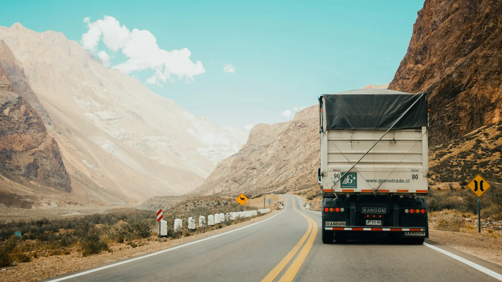 A cargo truck on a highway winding through dry mountains under a blue sky.