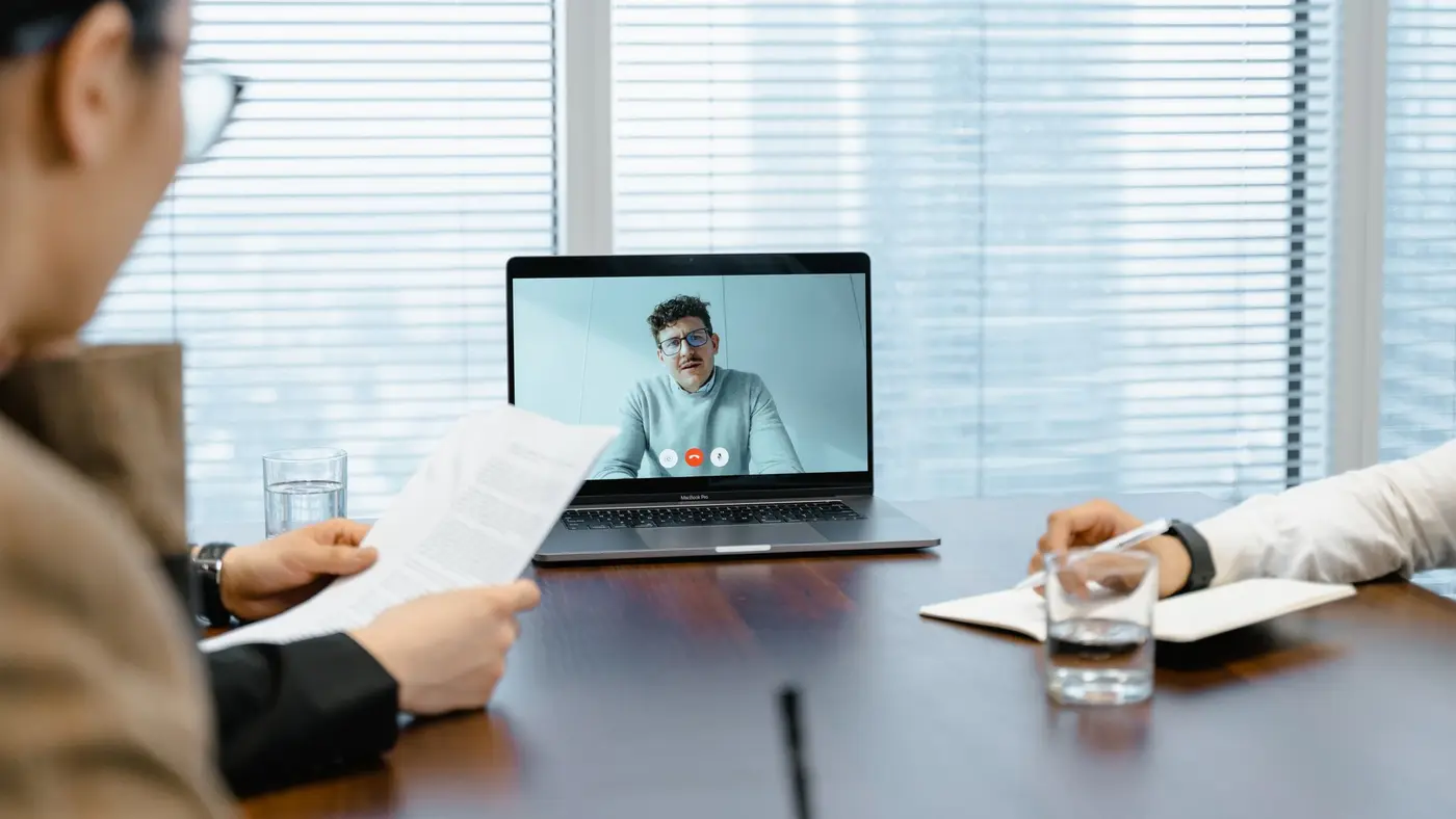 Two people at a meeting table reading notes while a third person joins them on a laptop video call.
