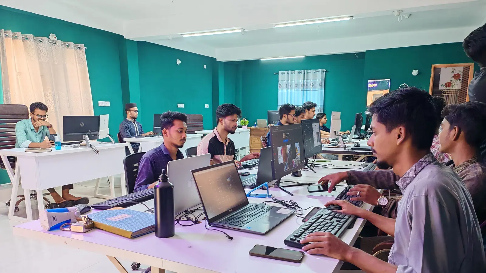 An open-plan office with teal walls, where a team of developers works at laptops and monitors along a long white desk.
