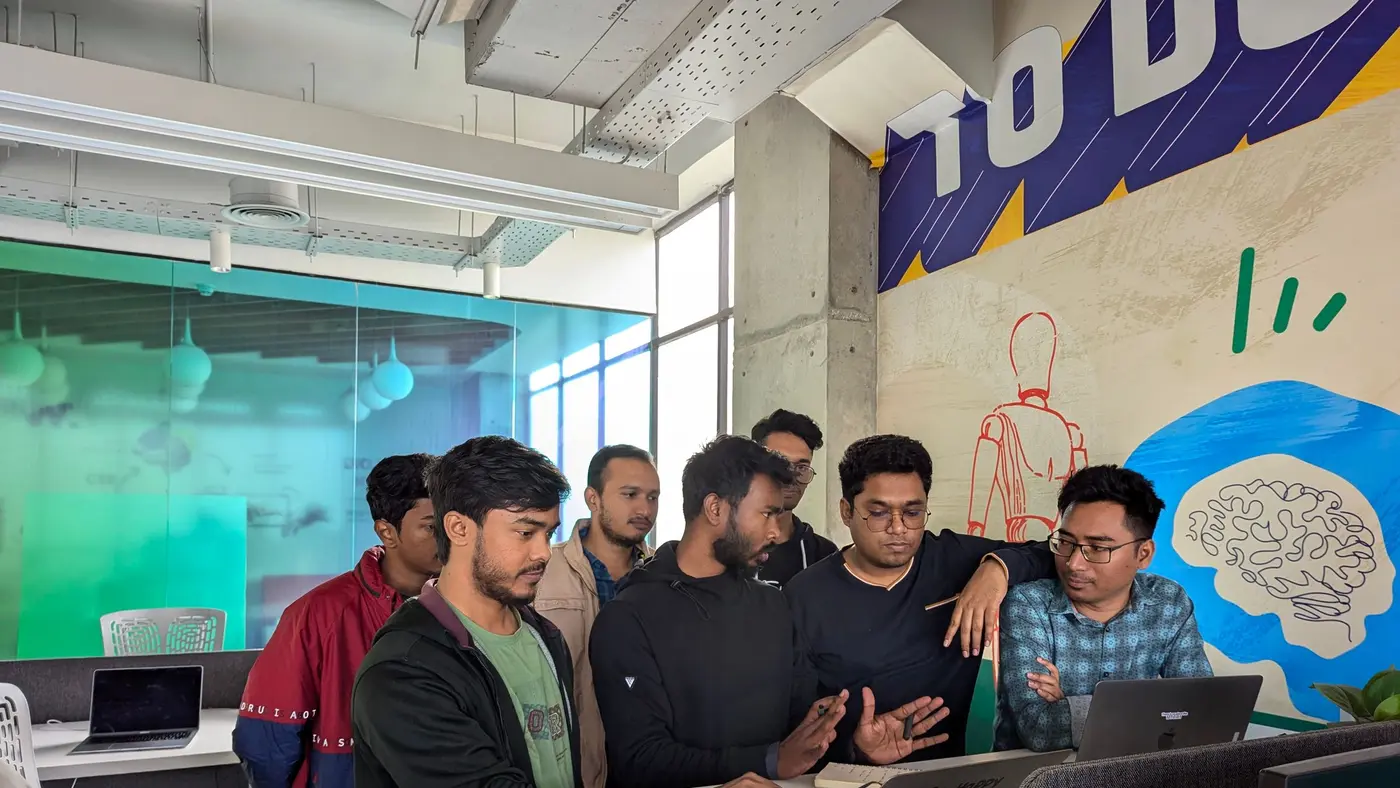 Six colleagues gathered around a laptop in a modern office, one gesturing at the screen while the others watch.