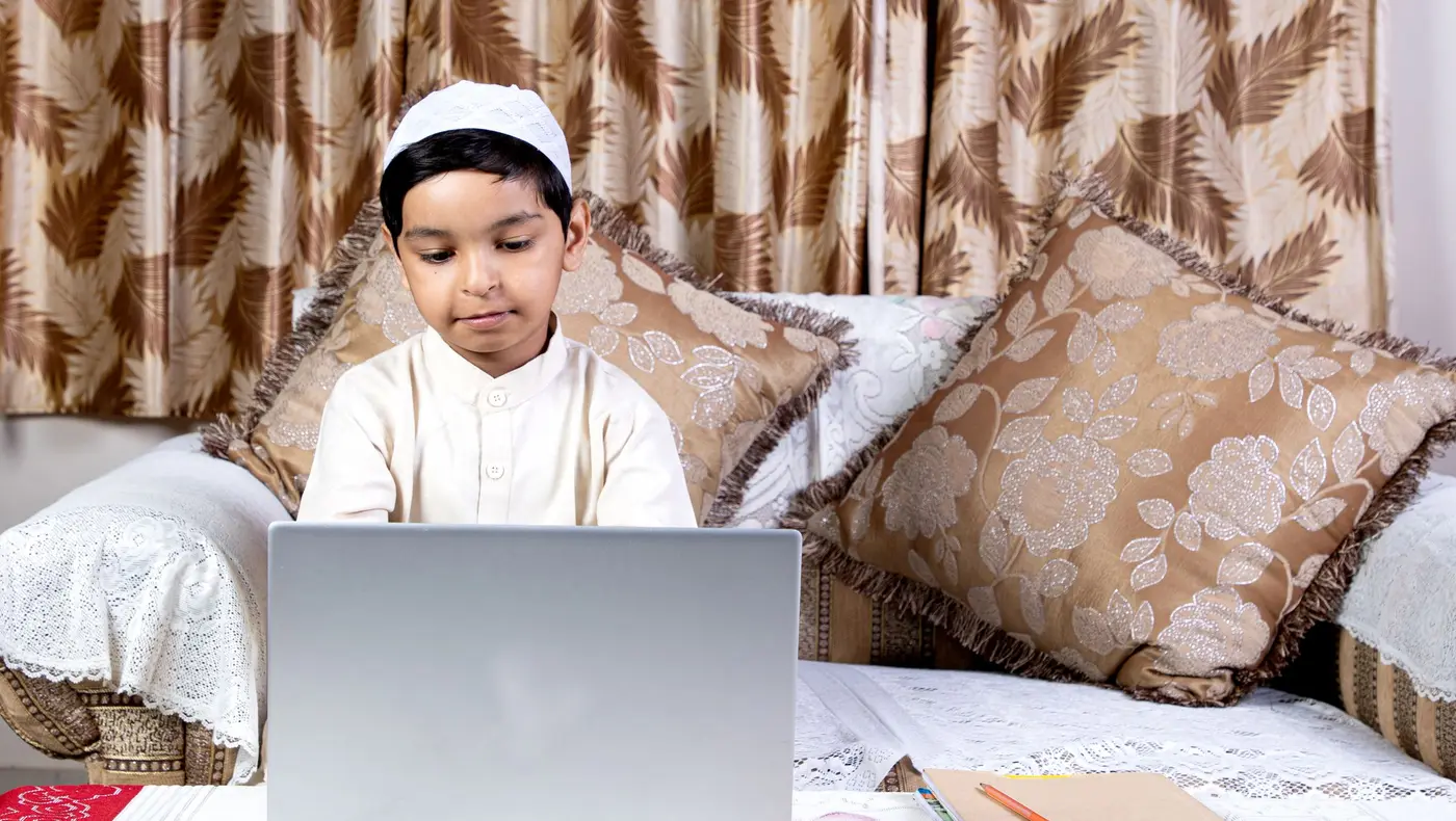 A young boy in a white kurta and cap attending an online class on a laptop at home