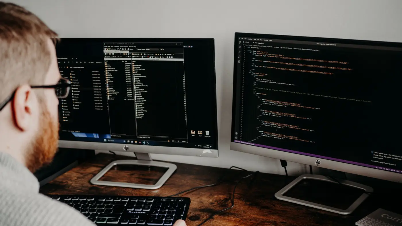 A developer at a wooden desk with two monitors, one showing a file manager and the other a code editor.