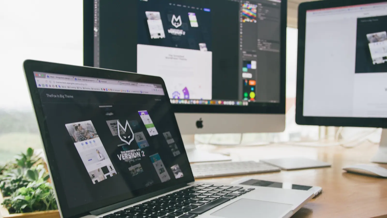 A desk with a laptop and two monitors, the laptop showing a website theme's demo page and the monitor showing the same design open in an image editor.