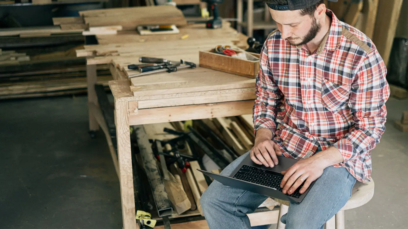 A carpenter sitting in a wood workshop, working on a laptop, surrounded by timber and tools.