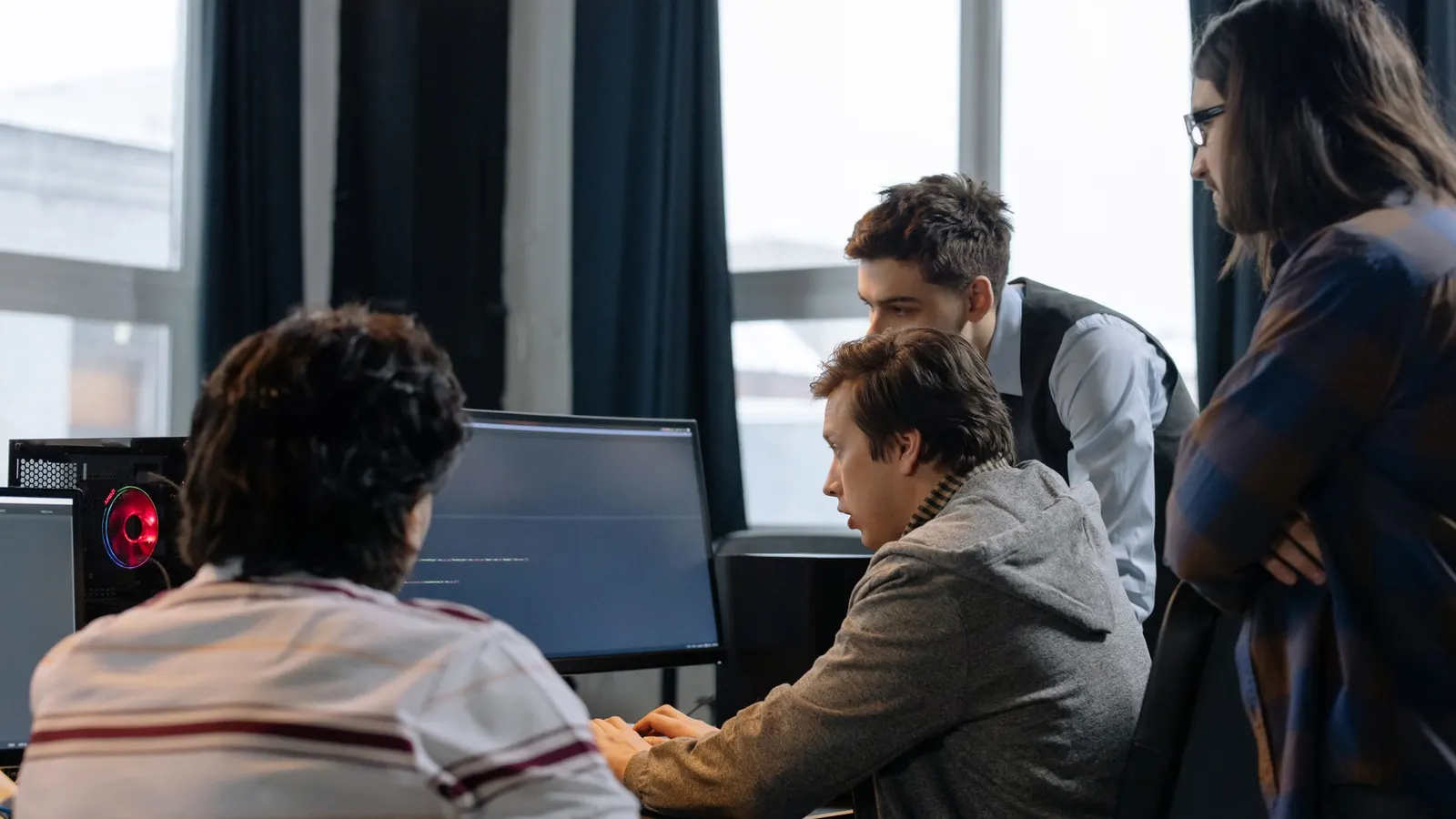 Four developers gathered around a monitor showing code, one typing and the others reading.