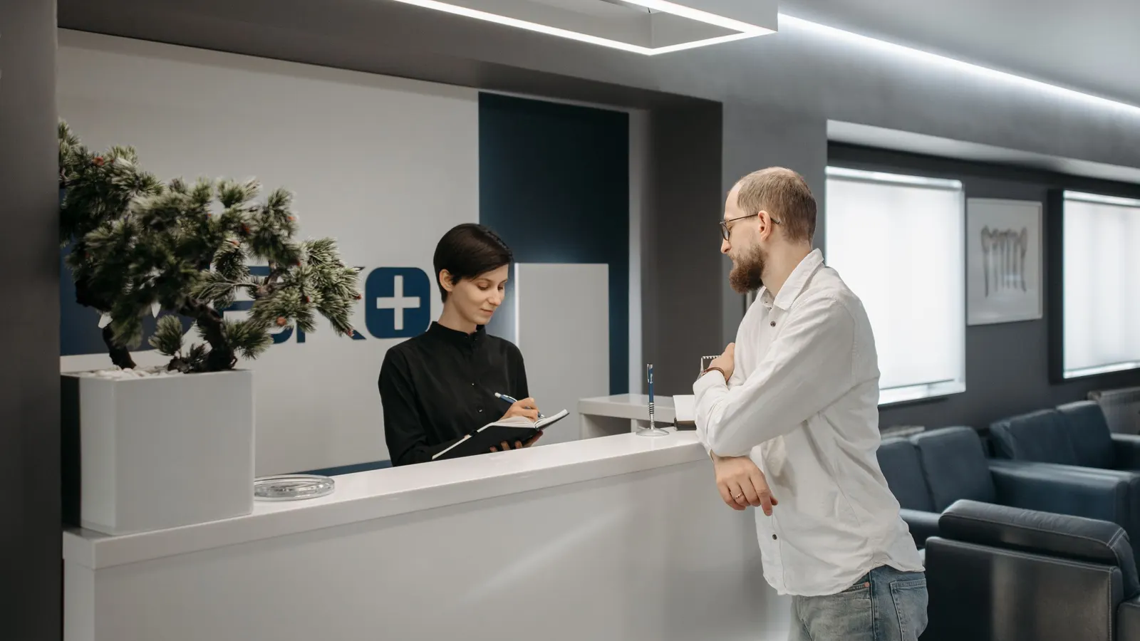 A receptionist behind a clinic front desk taking notes while a patient stands and talks to her.