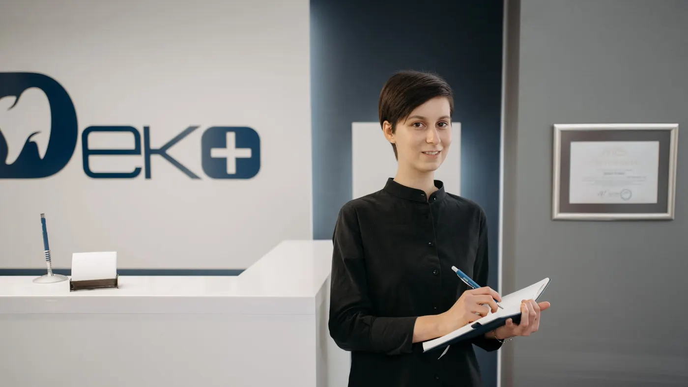 A receptionist in black standing at a dental clinic front desk with a notebook and pen.
