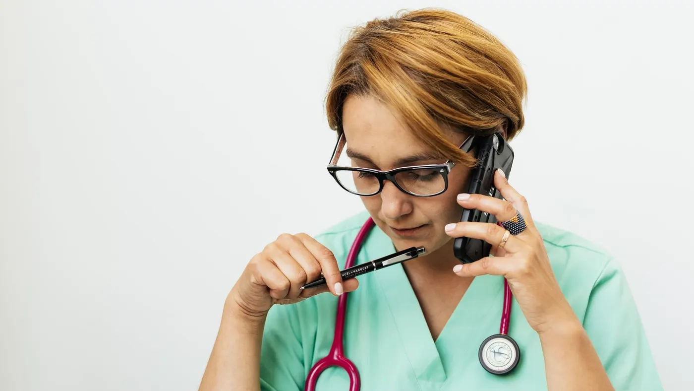 A nurse in green scrubs holding a phone to her ear and a pen in her hand.