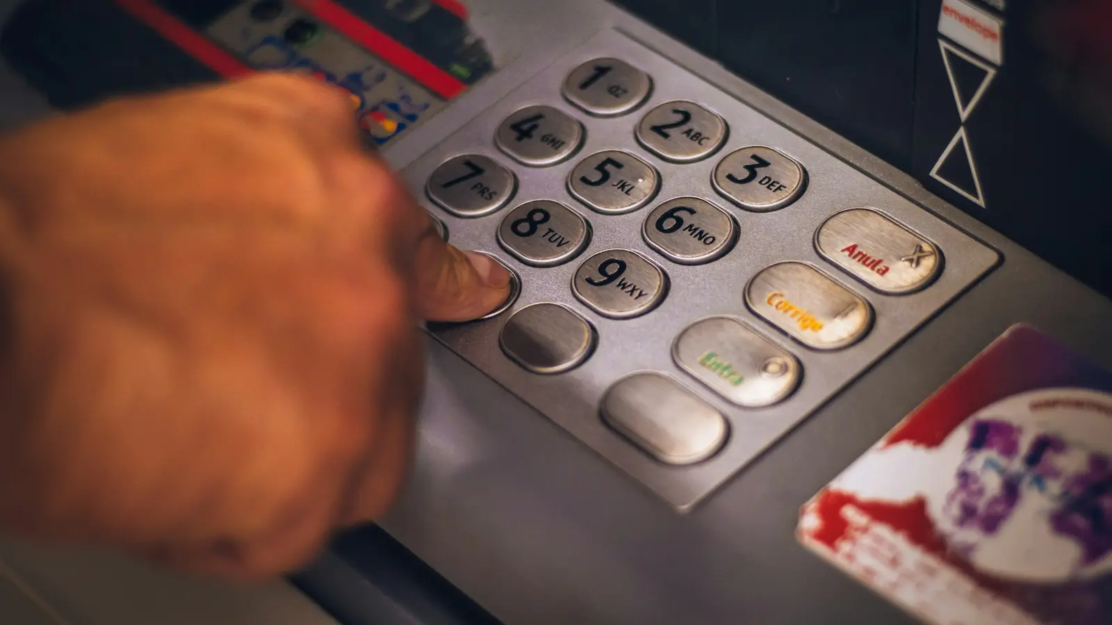 A hand pressing the keypad of an ATM, holding a red bank card.