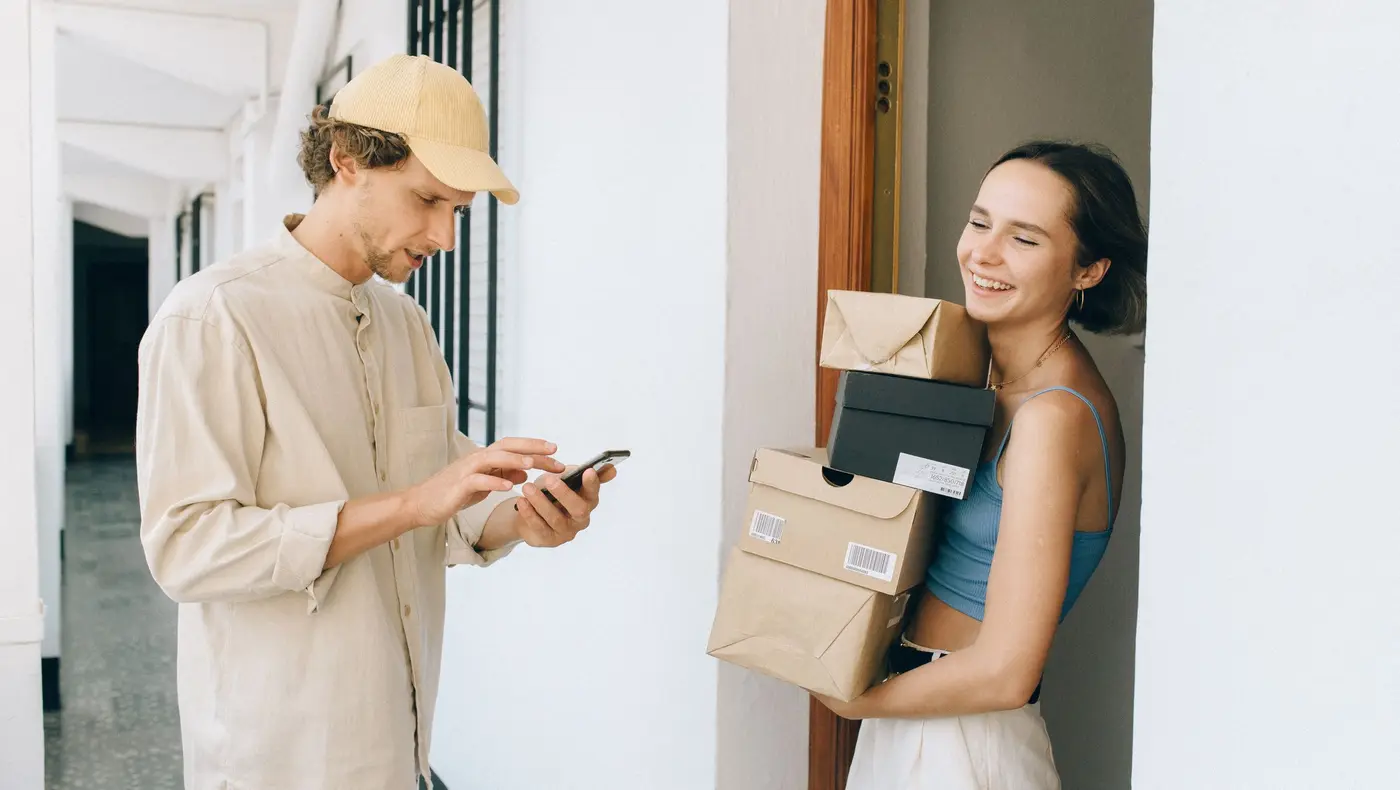 A courier checking his phone at a doorway while a customer holds a stack of delivered parcels