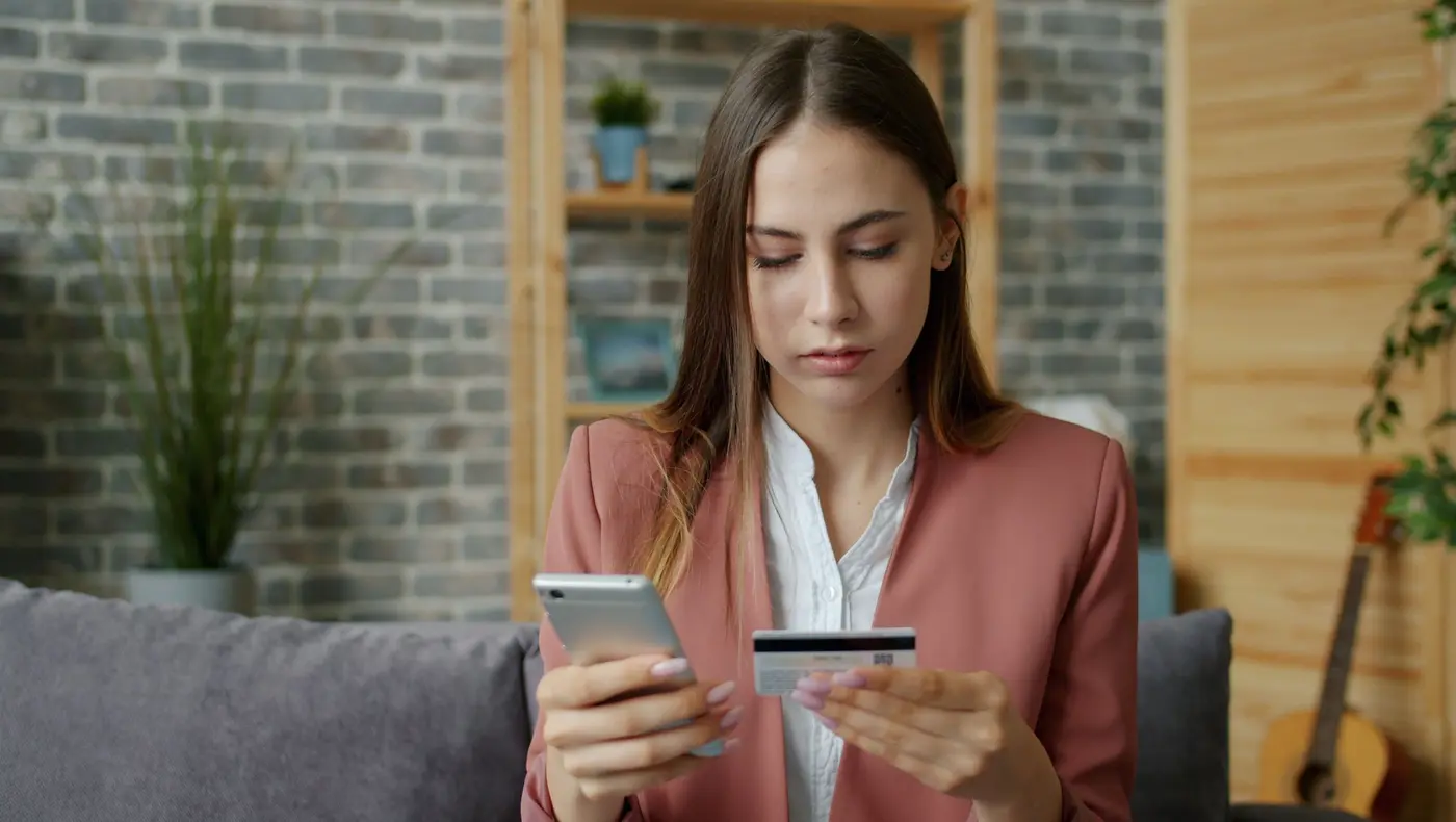 A woman on a sofa paying online, holding a smartphone in one hand and a bank card in the other