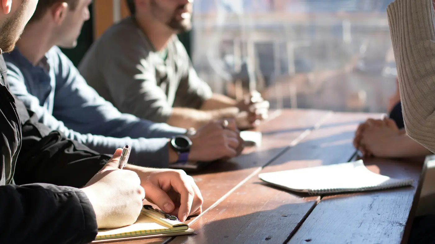 Two people talking across a wooden table with notebooks and papers.