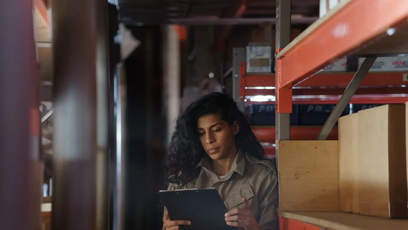 A warehouse worker checking stock on a clipboard between racks of boxes