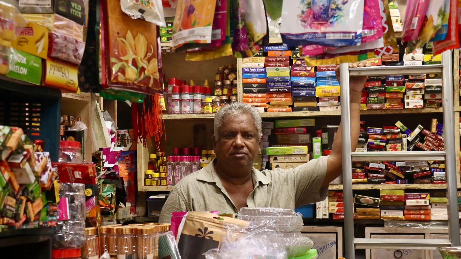 A shopkeeper at the counter of a small Indian store, surrounded by packed shelves of stock