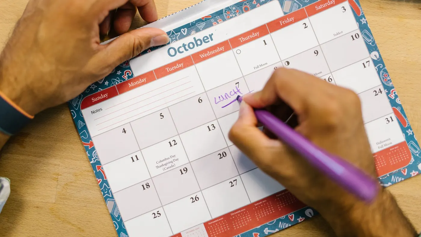 A hand writing on a paper wall calendar for October with a purple pen.