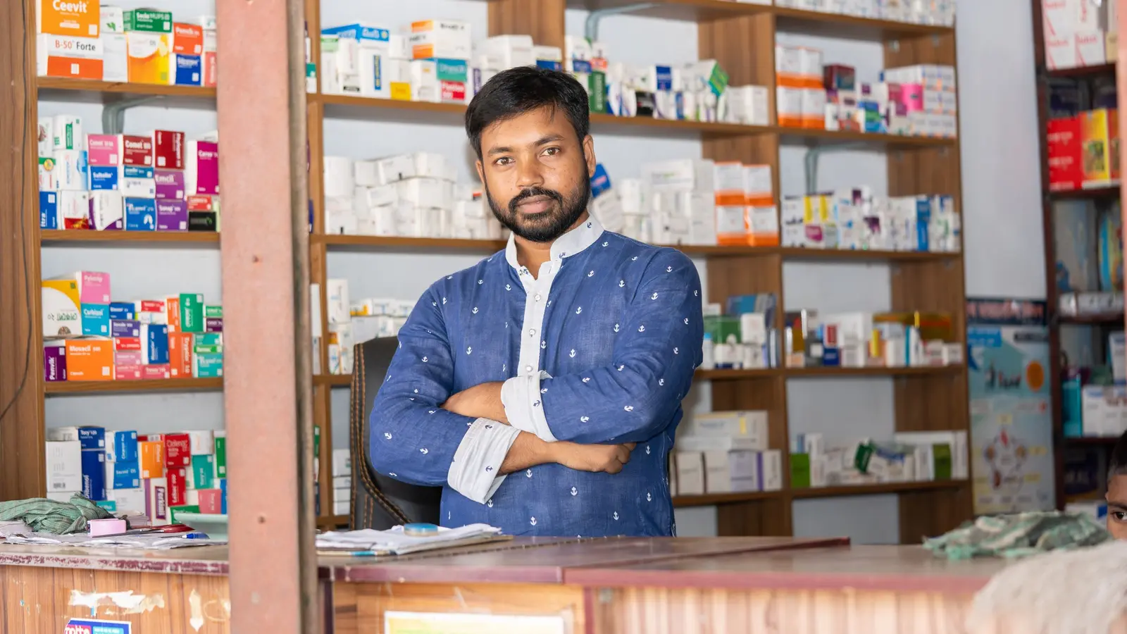 A chemist standing with folded arms behind the counter of a small medical store, shelves of medicine boxes behind him