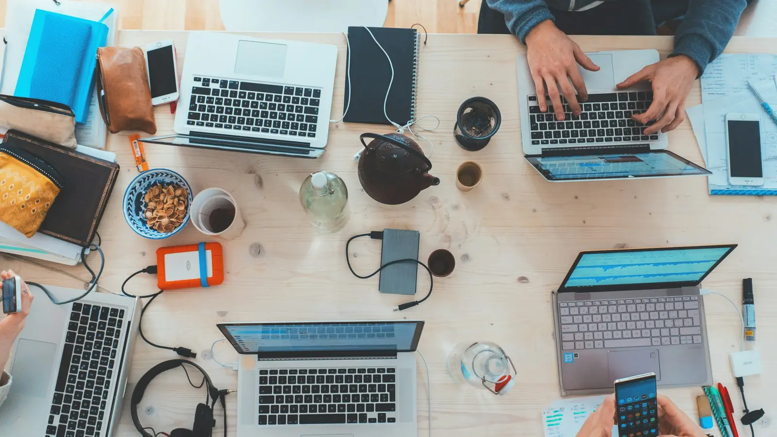 A desk from above covered with laptops, notebooks, phones and coffee cups as several people work.