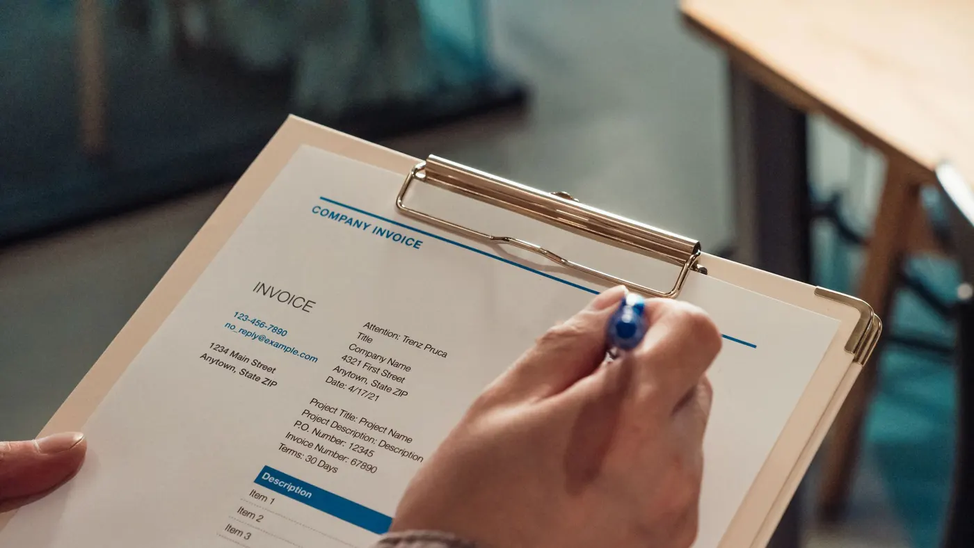 A hand holding a pen over a printed company invoice on a clipboard.