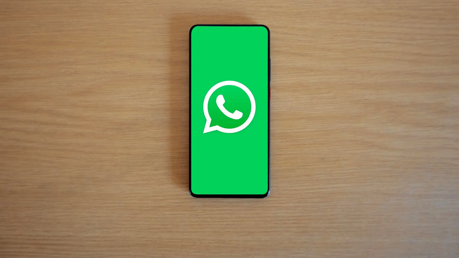 A smartphone lying on a wooden table with the WhatsApp logo on its screen, seen from above.