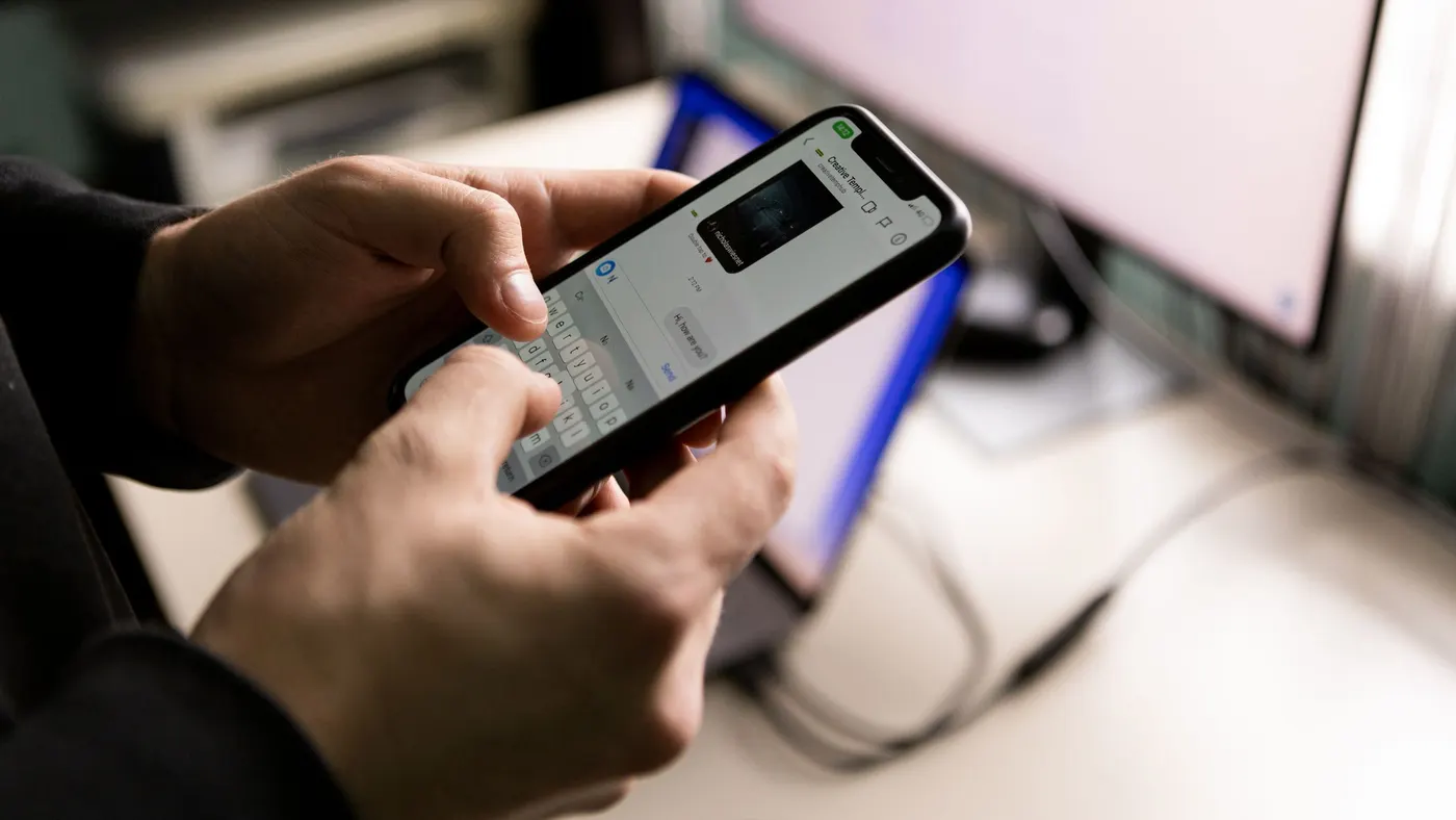 Two hands typing a reply in a chat app on a smartphone, with a computer monitor blurred behind.