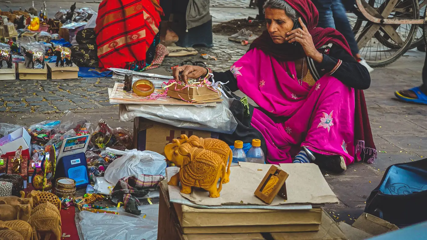 A street vendor in a pink sari talking on a phone, seated among wooden elephants and handicrafts at a Jaipur market.